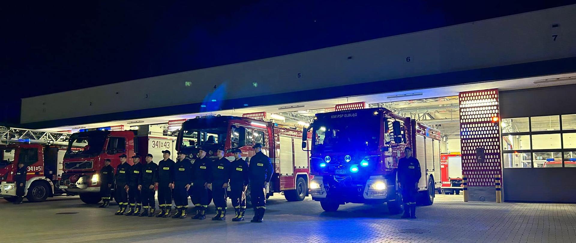Strażacy stoją w szeregu i oddają hołd swojemu koledze. Za nimi stoją pojazdy pożarnicze z włączonymi światłami alarmowymi. W tle widoczny budynek remizy strażackiej.