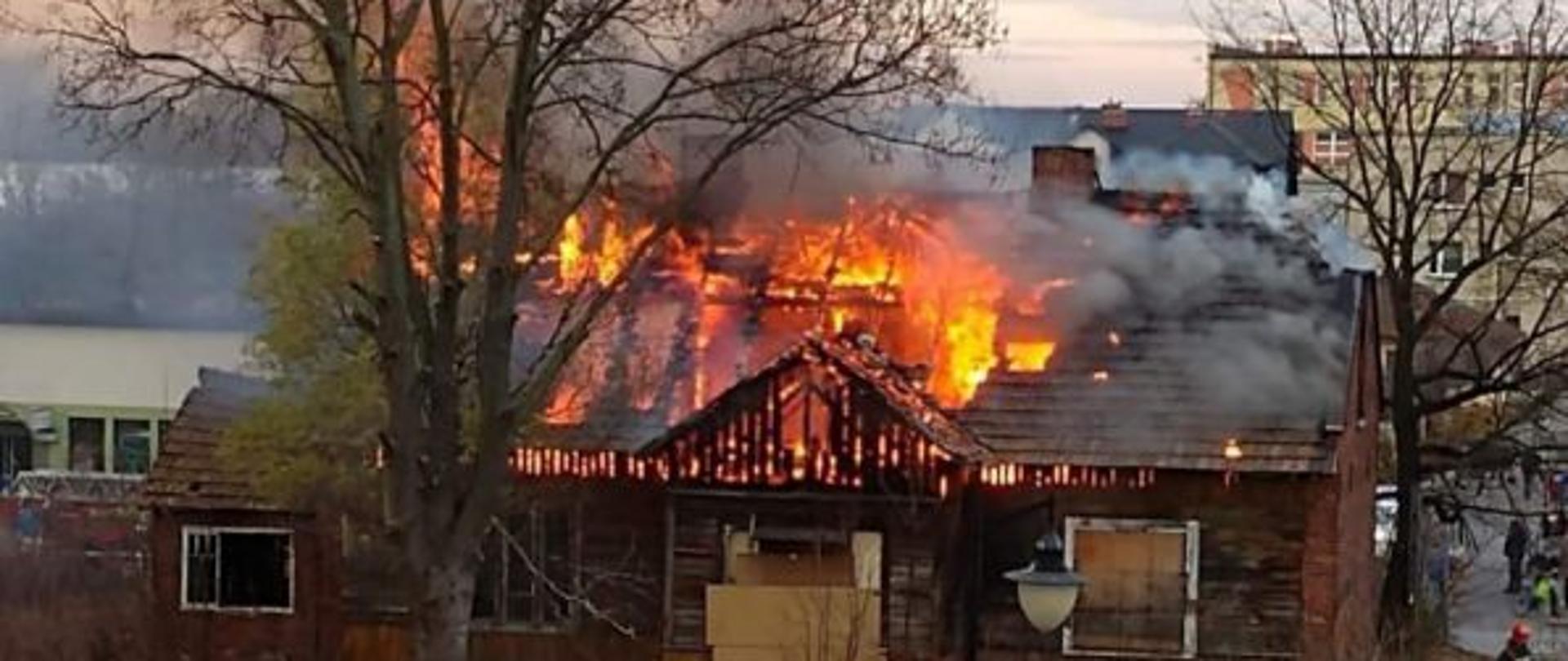 Na zdjęciu widoczny budynek z płonącym poddaszem i dachem. Budynek parterowy z zastawionymi otworami okiennymi i drzwiowymi. Nad budynkiem unoszą się chmury dymu pożarowego.