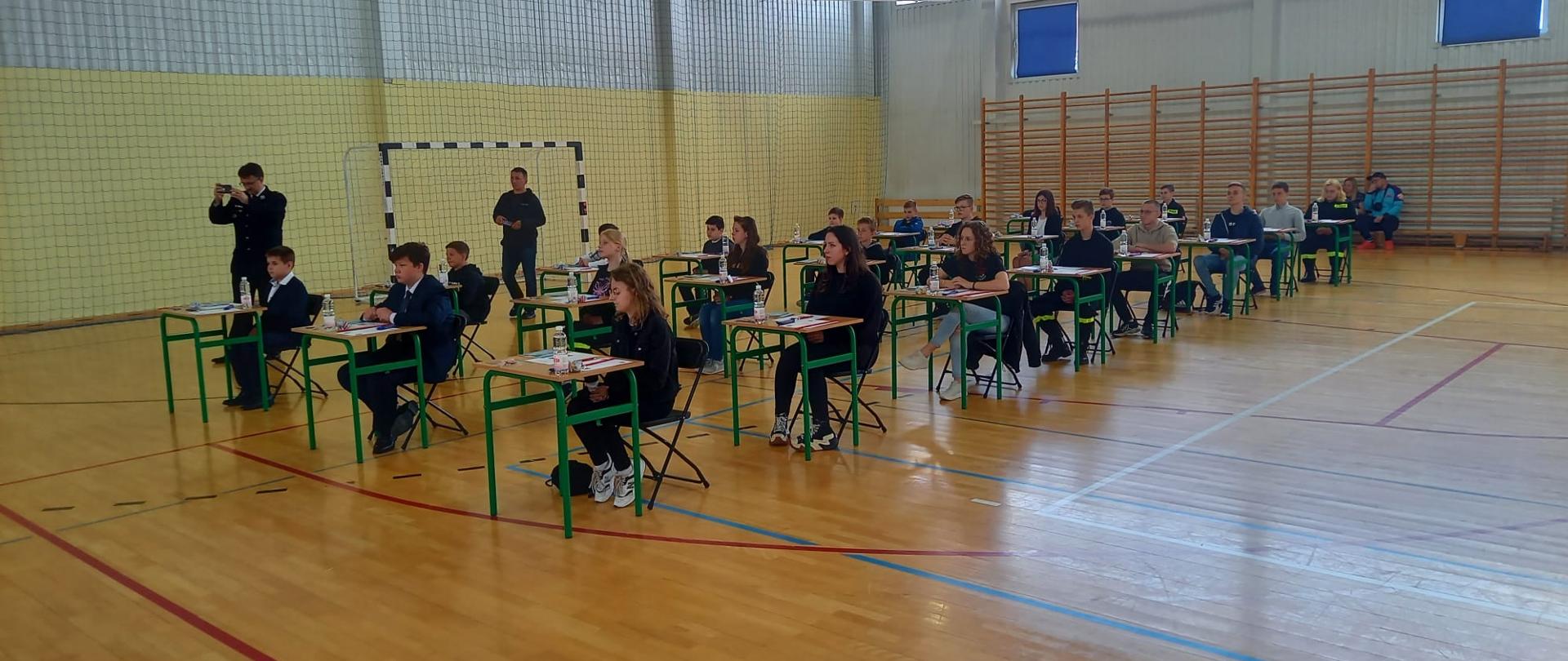 uczestnicy konkursu siedzą przy stolikach ustawionych na hali sportowej
