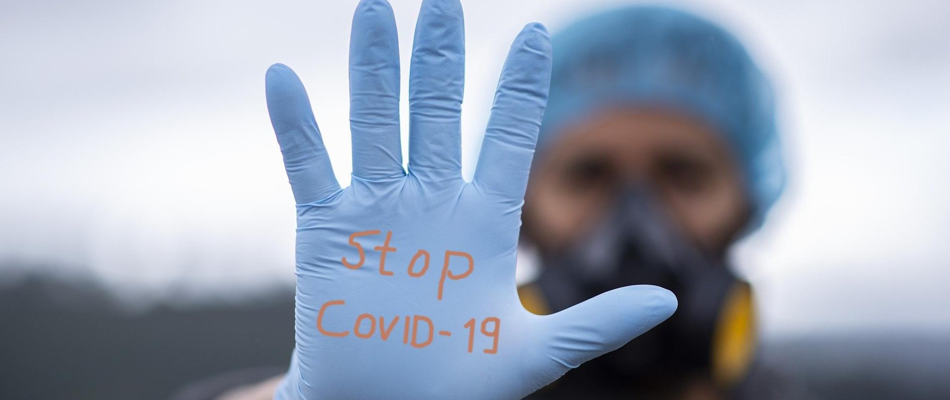 W tle człowiek w masce ochronnej i czepku, na pierwszym planie jego wyciągnięta dłoń w rękawiczce ochronnej z napisem Stop Covid-19