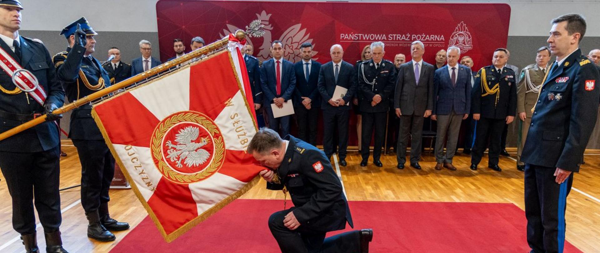 St. bryg. Paweł Kielar Opolski Komendant Wojewódzki Państwowej Straży Pożarnej oddaje honor sztandarowi Komendy Wojewódzkiej PSP w Opolu klęcząc na jedno kolano na czerwonym dywanie. Za oddającym honory stoi Komendant Główny Państwowej Straży Pożarnej nadbryg. dr inż. Mariusz Feltynowski. W tle stoją funkcjonariusze w stanie spoczynku, zaproszeni goście i przedstawiciele służb i instytucji współpracujących z Państwową Strażą Pożarną stoją na tle banneru koloru czerwonego z białym logo PSP, godłem Polski oraz napisem Państwowa Straż Pożarna Komenda Wojewódzka PSP w Opolu. Uroczystość odbywa się na sali gimnastycznej Komendy Wojewódzkiej PSP w Opolu.