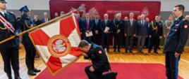 St. bryg. Paweł Kielar Opolski Komendant Wojewódzki Państwowej Straży Pożarnej oddaje honor sztandarowi Komendy Wojewódzkiej PSP w Opolu klęcząc na jedno kolano na czerwonym dywanie. Za oddającym honory stoi Komendant Główny Państwowej Straży Pożarnej nadbryg. dr inż. Mariusz Feltynowski. W tle stoją funkcjonariusze w stanie spoczynku, zaproszeni goście i przedstawiciele służb i instytucji współpracujących z Państwową Strażą Pożarną stoją na tle banneru koloru czerwonego z białym logo PSP, godłem Polski oraz napisem Państwowa Straż Pożarna Komenda Wojewódzka PSP w Opolu. Uroczystość odbywa się na sali gimnastycznej Komendy Wojewódzkiej PSP w Opolu.