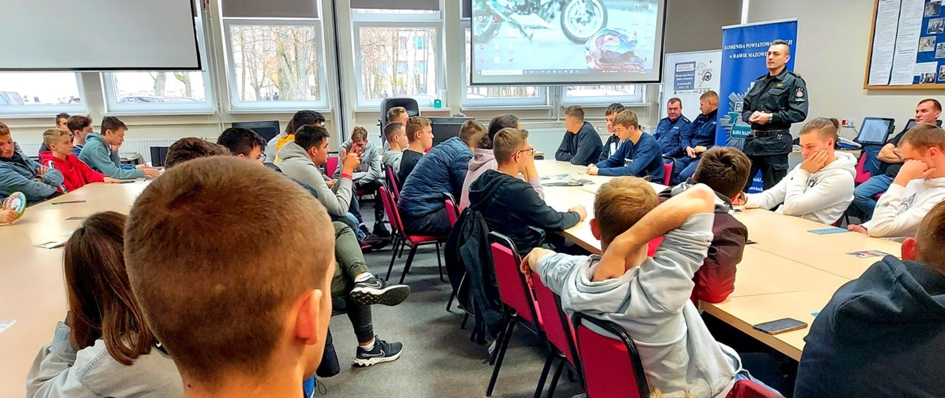 W sali szkolnej przy złączonych ławkach siedzą uczniowie, a przed nim znajdują się dwaj policjanci oraz jeden strażak, którzy prowadzą debatę na temat bezpieczeństwa.