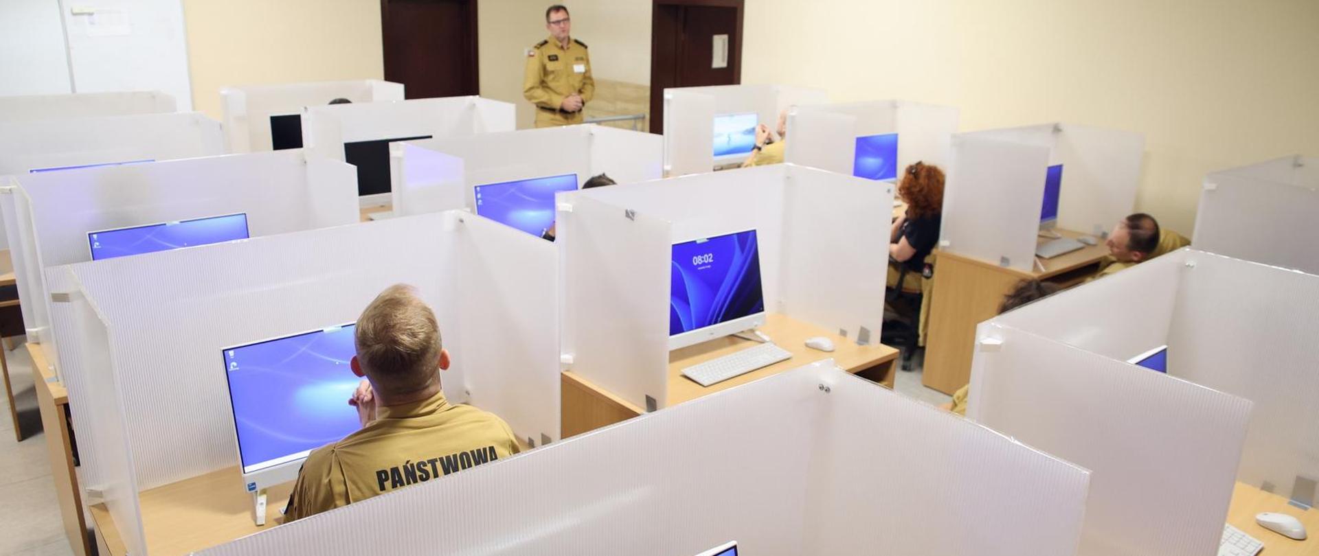 Sala egzaminacyjna, przy biurkach na których stoi komputer oraz osłona siedzą uczestnicy egzaminu, w głębi po prawej stojący funkcjonariusz CS PSP