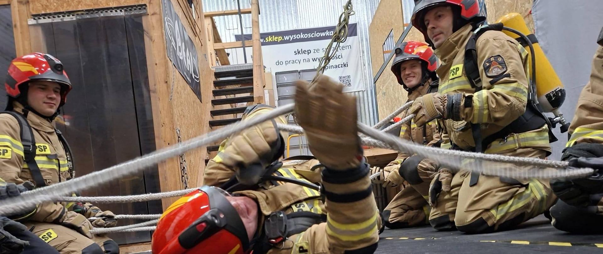 Czterech strażaków w mundurach i czerwonych hełmach podczas szkolenia.
