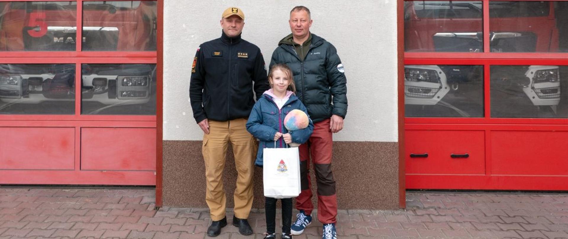 Na zdjęciu widać oficera z Państwowej Straży Pożarnej oraz dziewczynkę z Tatą przed garażami JRG Ustrzyki Dolne.