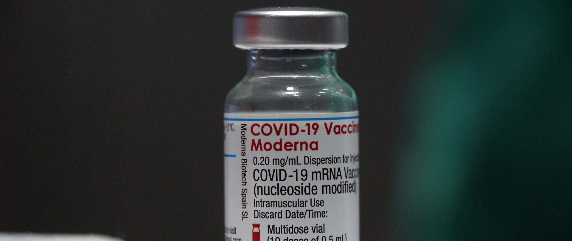 Na zdjęciu fiolka ze szczepionką Moderna. Fiolka stoi na stole. Jest przeźroczysta, szklana. Na fiolce naklejka, na której jest napis Covid19 - Moderna. Tło ciemne. 