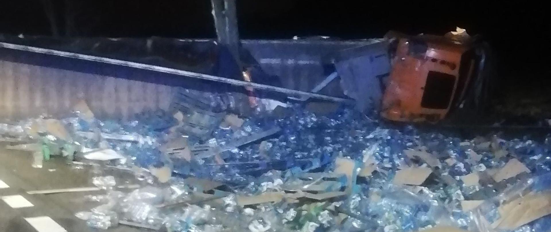 Zdjęcie przewróconej ciężarówki na bok. Na jezdni porozrzucana zawartosc naczepy - butelki z wodą.