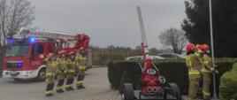 Zdjęcie przedstawia strażaków PSP, którzy z okazji Dnia Flagi Rzeczypospolitej Polskiej podnoszą flagę Polski na maszt. Na zdjęciu po lewej stronie widoczny jest samochód pożarniczy - podnośnik hydrauliczny z włączonymi niebieskimi światłami alarmowymi. Przed pojazdem stoi 4 strażaków, pierwszy z nich salutuje. W środkowej części zdjęcia widać działko wodno - pianowe. W prawej części zdjęcia znajduje się 3 strażaków, którzy stoją przed maszem i podnoszą flagę Polski na maszt.