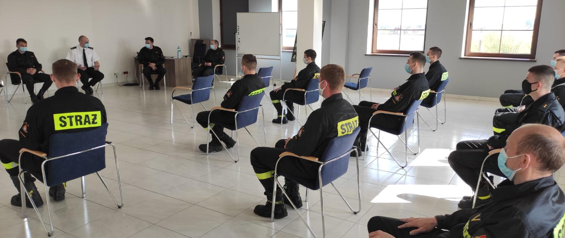 Na zdjęciu widać strażaków siedzących na krzesłach w pomieszczeniu. W tle widać okna. Wszyscy strażacy mają ubrane maseczki zasłaniające usta i nos. 