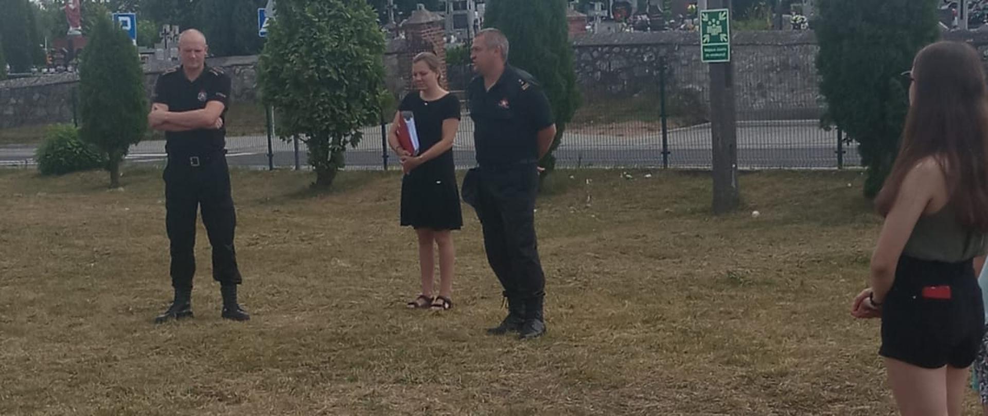 Dwóch strażaków w czarnych ubraniach na krótki rękaw stają na boisku przodem do obiektywu. Między nimi stoi kobieta. Z prawej strony zdjęcia bokiem również widoczna jest kobieta.