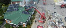 Widok obiektu objętego pożarem z lotu ptaka, widać podnośnik strażacki i strażaków działajcych na dachu budynku 