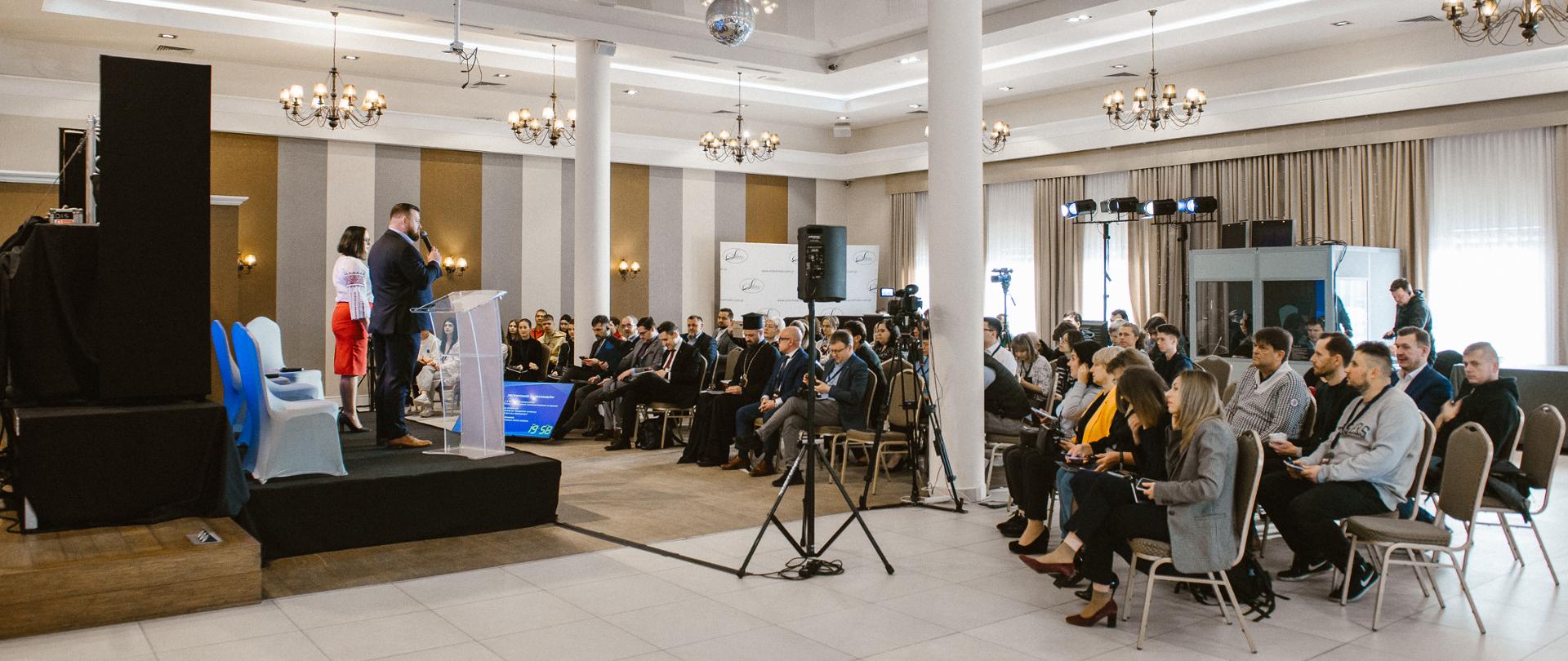 Na sali w kilku rzędach siedzą uczestnicy konferencji i słuchają mężczyzny i kobiety stojących na podium z krzesłami i mównicą