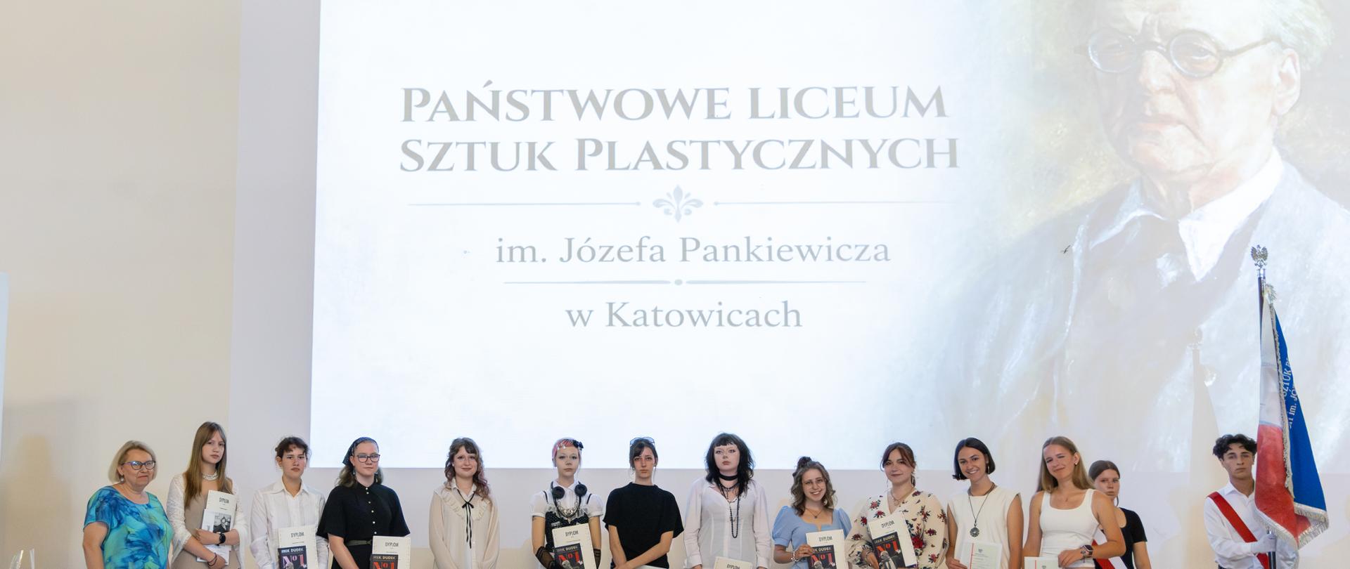 Fotografia z zakończenia roku szkolnego ukazująca osoby na tle wyświetlanego slajdu z nazwą szkoły i wizerunkiem patrona.