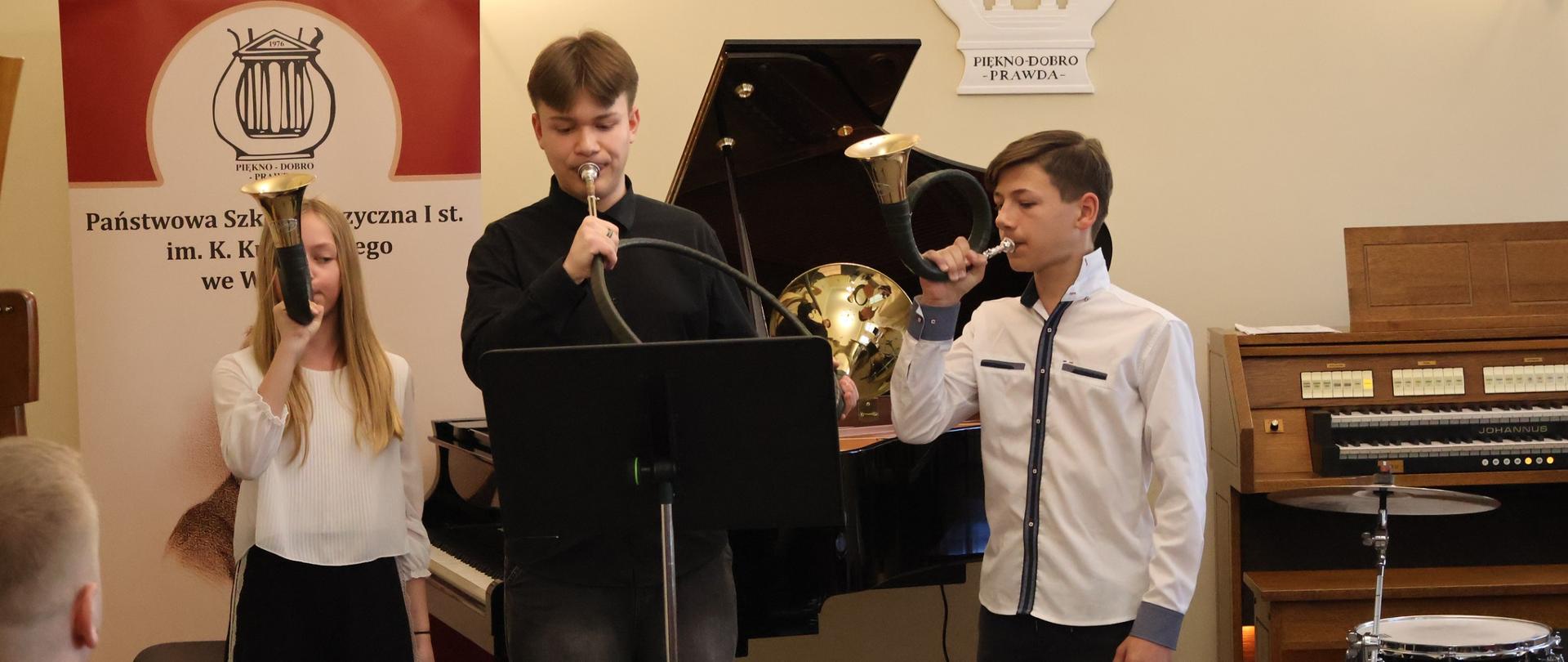 Troje uczniów grających na rogach myśliwskich w auli szkoły.