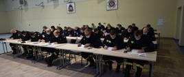 Grupa strażaków OSP w ubraniach koszarowych siedzi za stolikami i wypełnia ankietę. 