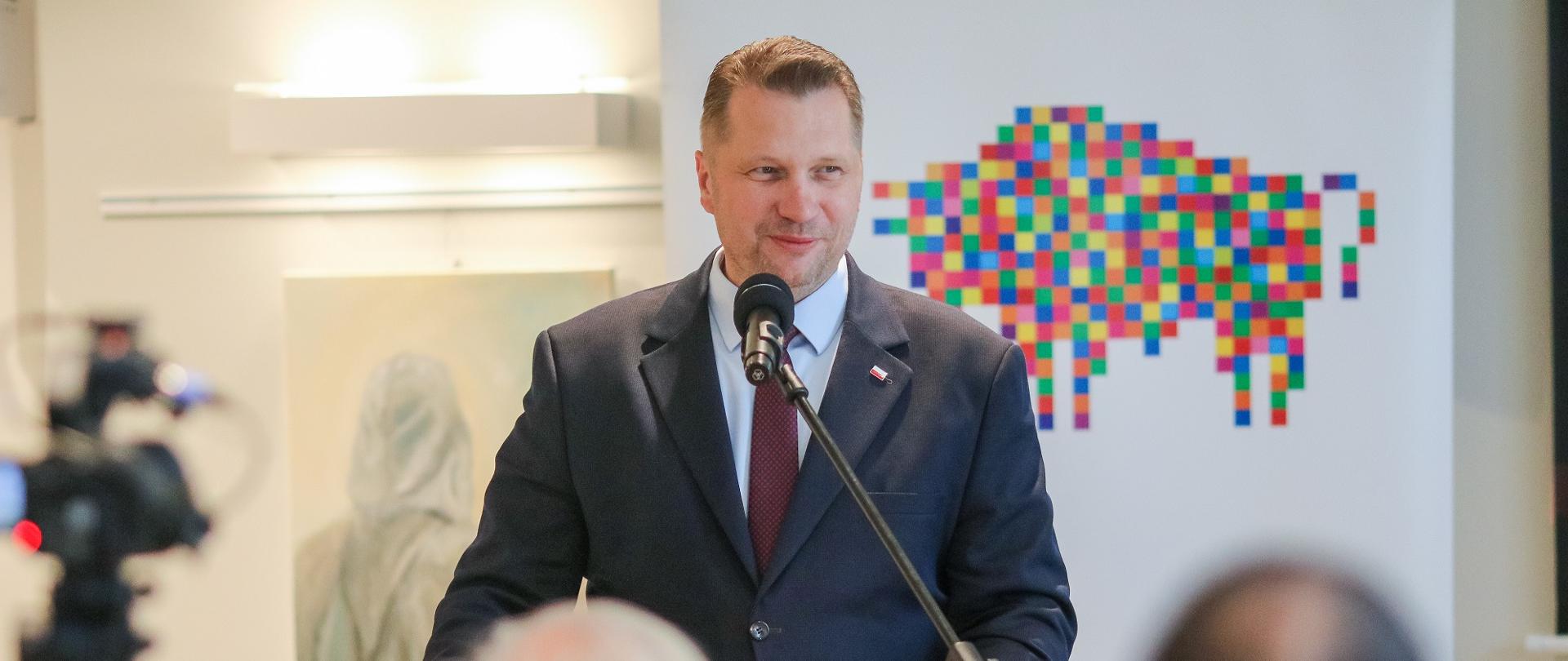 Minister Czarnek stoi i mówi do mikrofonu na stojaku, przed nim zarysy osób siedzących na sali, za nim baner w kolorowy geometryczny wzorek na białym tle.