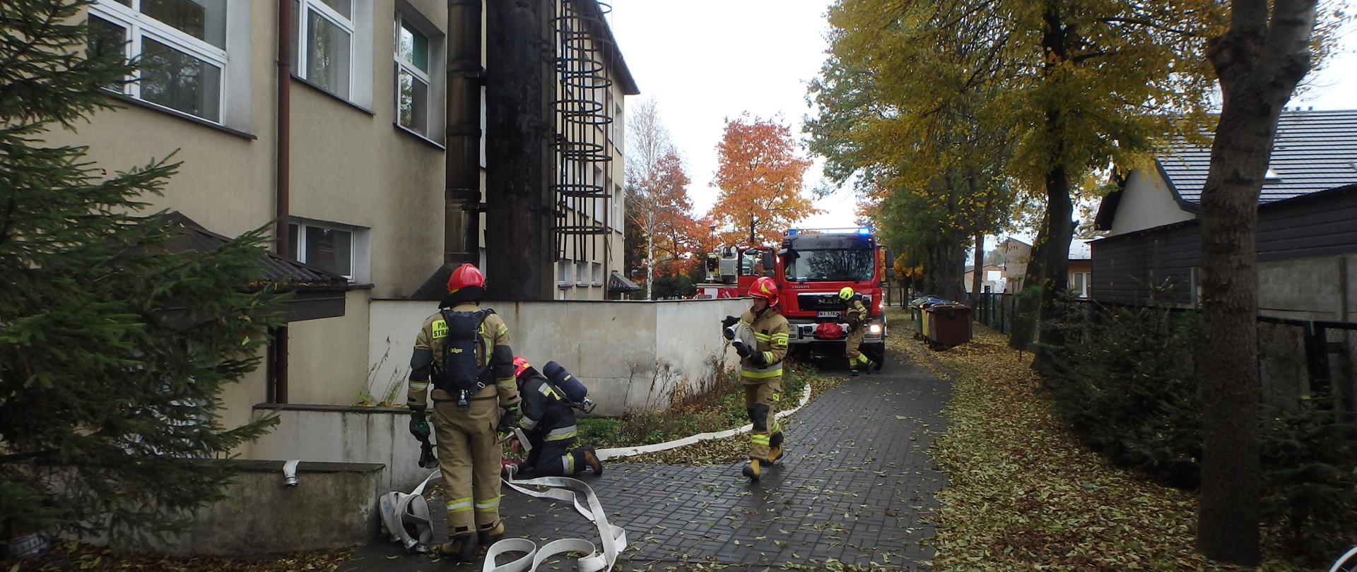 Przed budynkiem szkoły podstawowej w tle ustawione są samochody ratowniczo- gaśnicze. Przed wejściem trzech strażaków w pełnym umundurowaniu specjalnym wykonuje ćwiczenia ratowniczo- gasnicze.