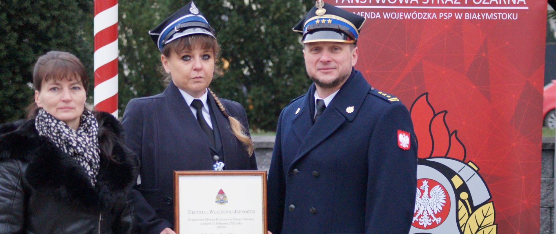 Na tle czerwonego banera stoją dwie kobiety i mężczyzna .Mężczyzna i jedna kobieta ubrani w mundur strażacki.