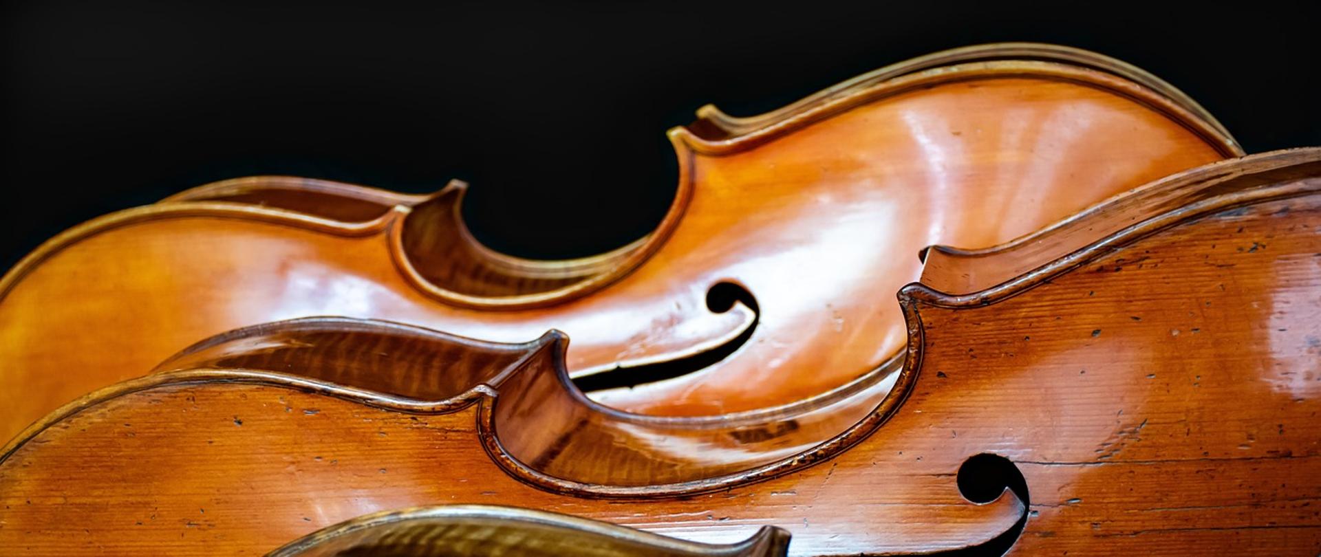 Zdjęcie pokazuje fragment trzech wiolonczeli na ciemnym tle 