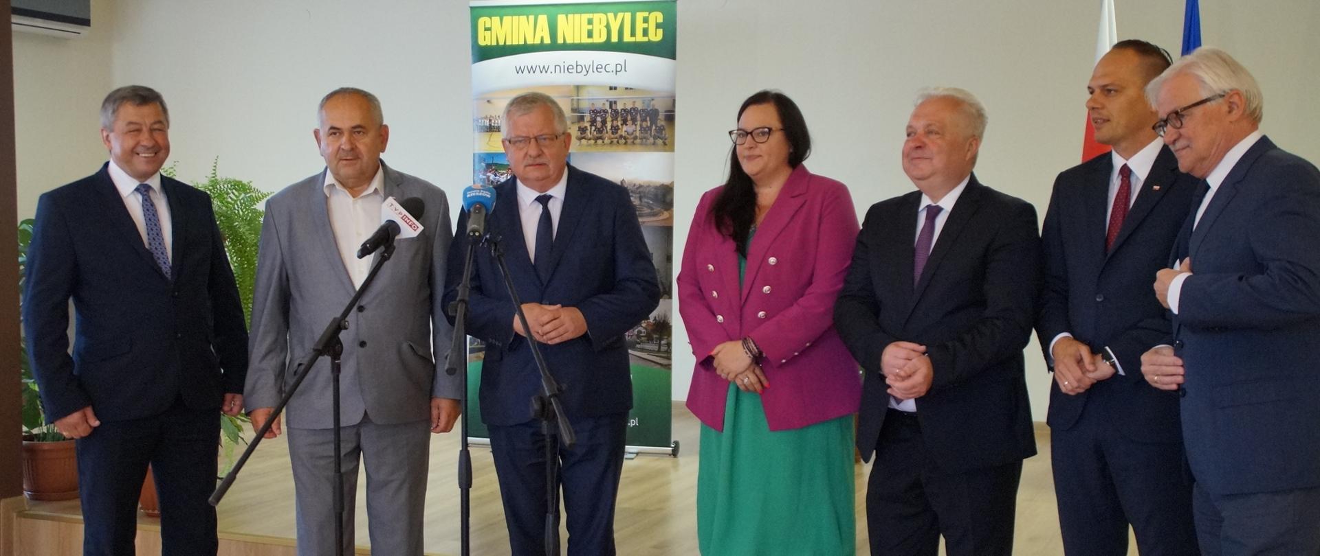 Grupa osób w sali stoi przed mikrofonami na statywach