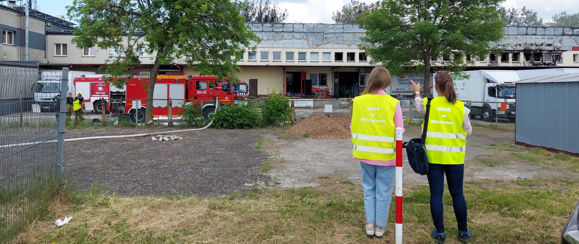 Dwie inspektorki WIOŚ w Warszawie stoją przed halą. Jedna pokazuje palcem w kierunku budynku. Przed halą stoi wóz strażacki oraz samochody dostawcze.