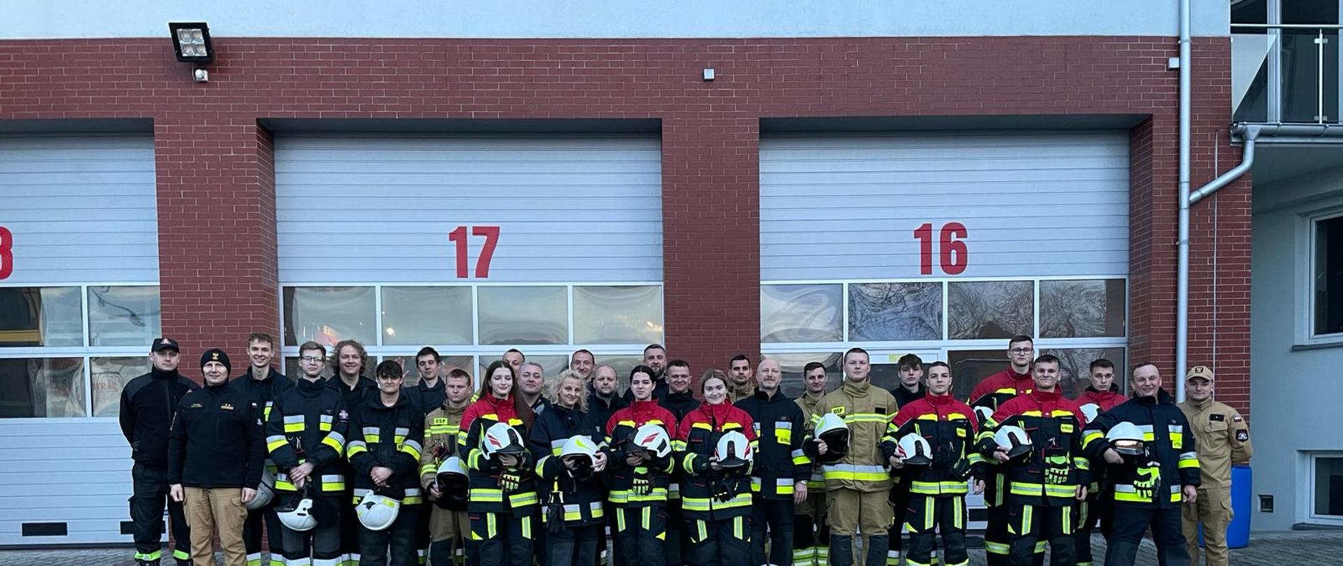 Kilkudziesięciu mężczyzn ubranych w czarne ubrania koszarowe strażackie z odblaskowymi paskami, czarnym butami stoją na polu przed bramami wyjazdowymi z garażu. W tle ściany z płytek w kolorze cegły oraz bramy z napisami