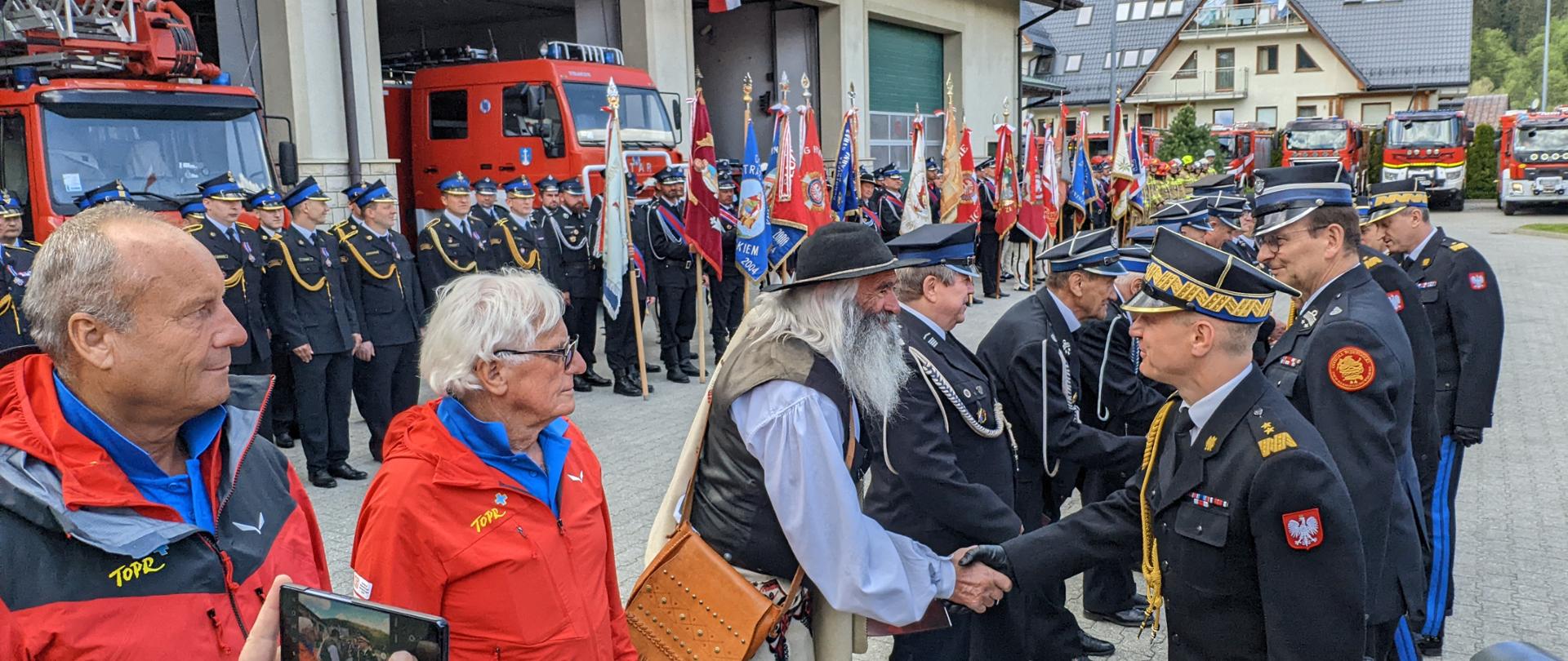 Komendant główny gratuluje zasłużonym osobom, które stoją w rzędzie. W tle poczty sztandarowe