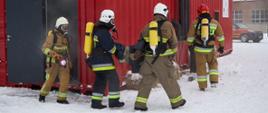 Czterech mężczyzn w kombinezonach, maskach i kaskach idzie obok trenażera; jeden trzyma w ręku latarkę, a dwóch wąż strażacki
