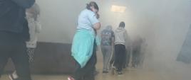 Grupa dzieci ewakuuje się gęsiego przez mocno zadymiony szkolny korytarz. Uczniowie idą pochyleni, trzymając dłonie lub ubrania przy twarzach, aby chronić drogi oddechowe przed dymem.