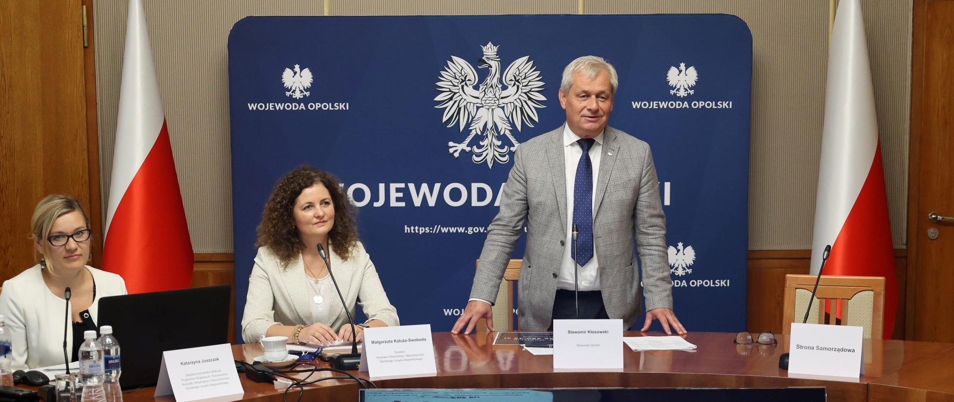 Na zdjęciu widoczni są za stołem: Wojewoda Opolski, Sławomir Kłosowski, Dyrektor Wydziału Infrastruktury i Nieruchomości w Opolu Małgorzata Kałuża-Swoboda wraz z zastępcą Katarzyną Juszczak. Przed stołem znajduje się wyświetlacz, na którym widnieje napis: "Rządowy Fundusz Rozwoju Dróg. Dofinansowanie na REMONTY na 2023 rok" oraz kwota 25289360 złotych i czterdzieści pięć groszy. W tle widoczna jest flaga Rzeczypospolitej Polskiej oraz ścianka z godłem państwowym i napisem Wojewoda Opolski.
