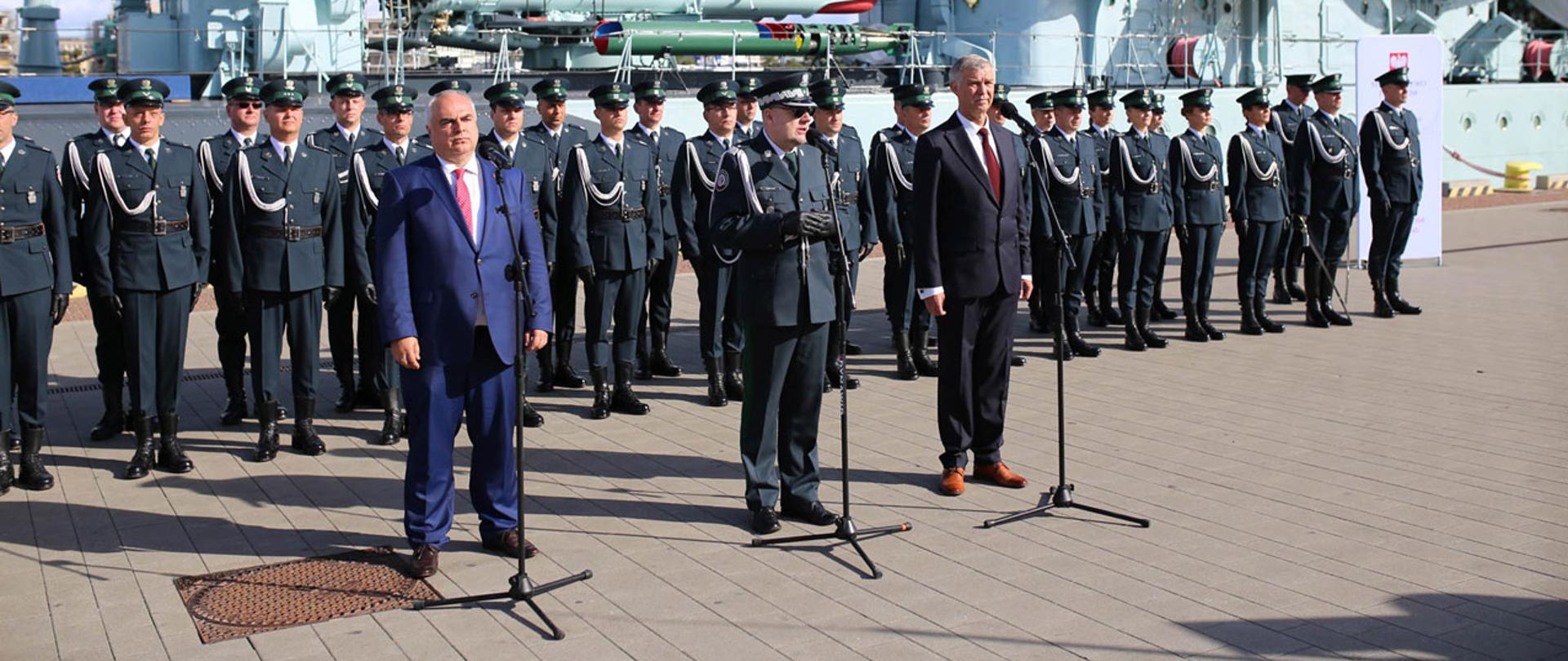 Od lewej: z-ca Szefa KAS Tomasz Słaboszowski, Szef KAS nadinsp. Piotr Walczak, dyrektor Izby Administracji Skarbowej w Gdańsku Leszek Synak, w tle funkcjonariusze KAS oraz okręt.