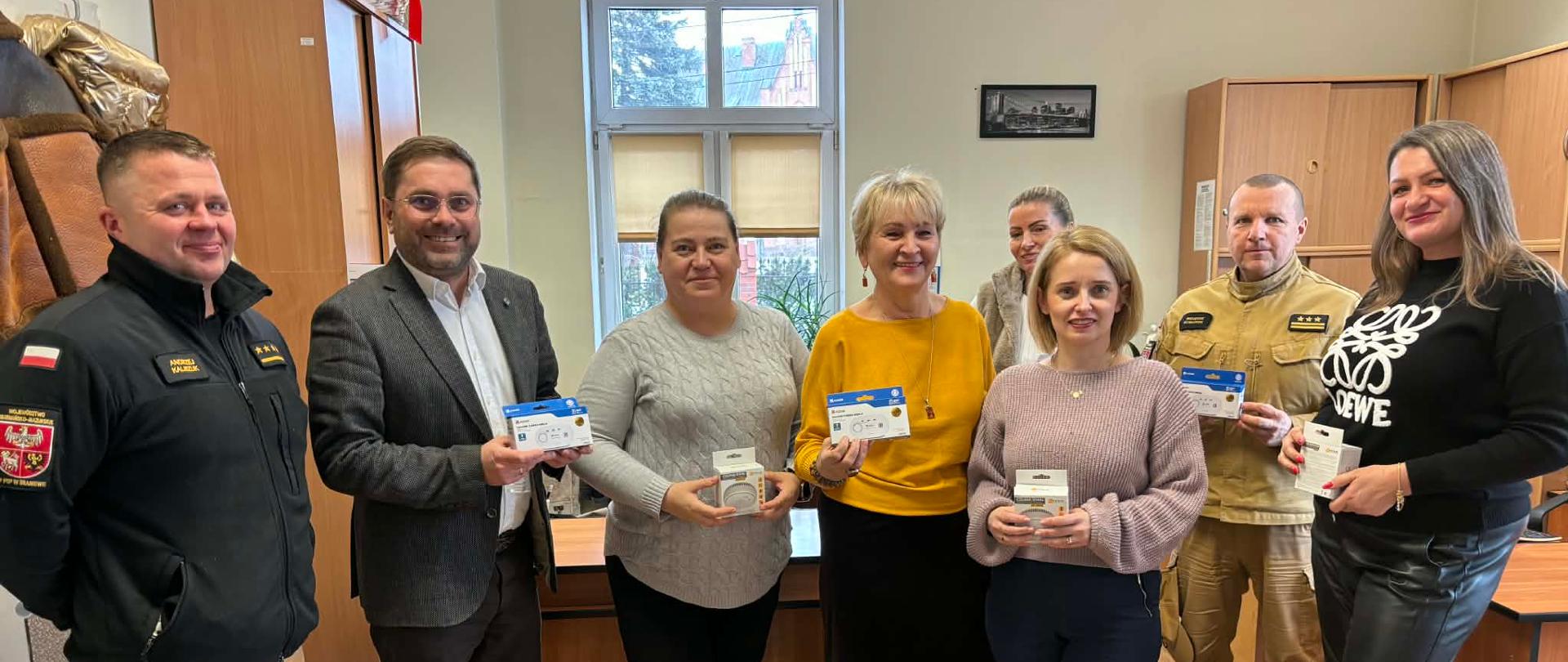 W gabinecie biura stoi pięć kobiet z wójtem gminy Braniewo oraz dwoma strażakami. Pozują do zdjęcia, trzymają w rekach czujki tlenku węgla.