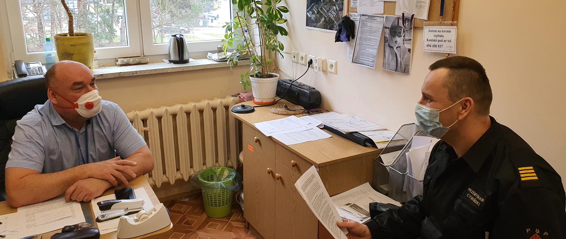 Zdjęcie przedstawia pomieszczenie biurowe, w którym siedzi dwóch mężczyzn. Po prawej strażak w czarnym mundurze przekazuje informacje mężczyźnie siedzącemu po lewej. Obaj mają założone maseczki ochronne. W tle okno, szafa i inne wyposażenie biurowe.