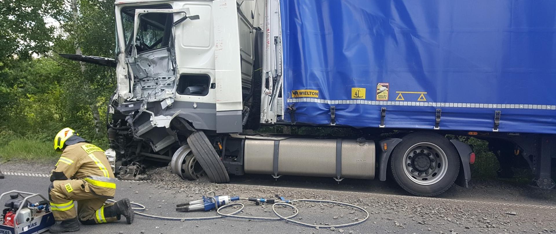 Dzień. Widok z boku na samochód ciężarowy z białą kabiną i niebieską plandeką na naczepie.Samochód stoi na poboczu szutrowym. Za nim i zboku widać drzewa. Zniszczona przednia części kabiny, drzwi kierowcy. Część przedniej osi wraz z felgą wyrwana z mocowań, częstościowy zagrzebana w szutrze pobocza. Przy pojeździe na asfalcie jezdni klęczy strażak ubrany w ubranie specjalne w kolorze piaskowym oraz hełm w kolorze żółtym. Strażak klęczy przy pompie hydraulicznej, z elementami w kolorze niebieskim. Do pompy za pomocą przewodów w kolorze szarym i niebieskim podłączony jest rozpieracz ramieniowy.
