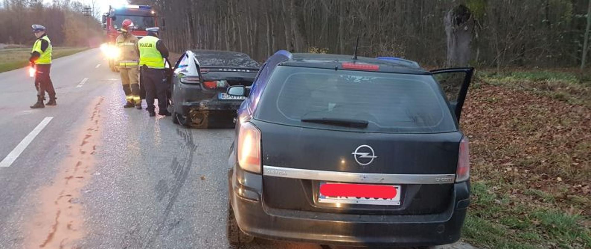 Przez środek zdjęcia biegnie droga. Pierwszy plan to auto osobowe, kombi, widoczne od tyłu. Przed nim stoi drugie auto z uszkodzonym tyłem. Najdalej przy górze zdjęcia widoczni strażacy i policjanci kierujący ruchem