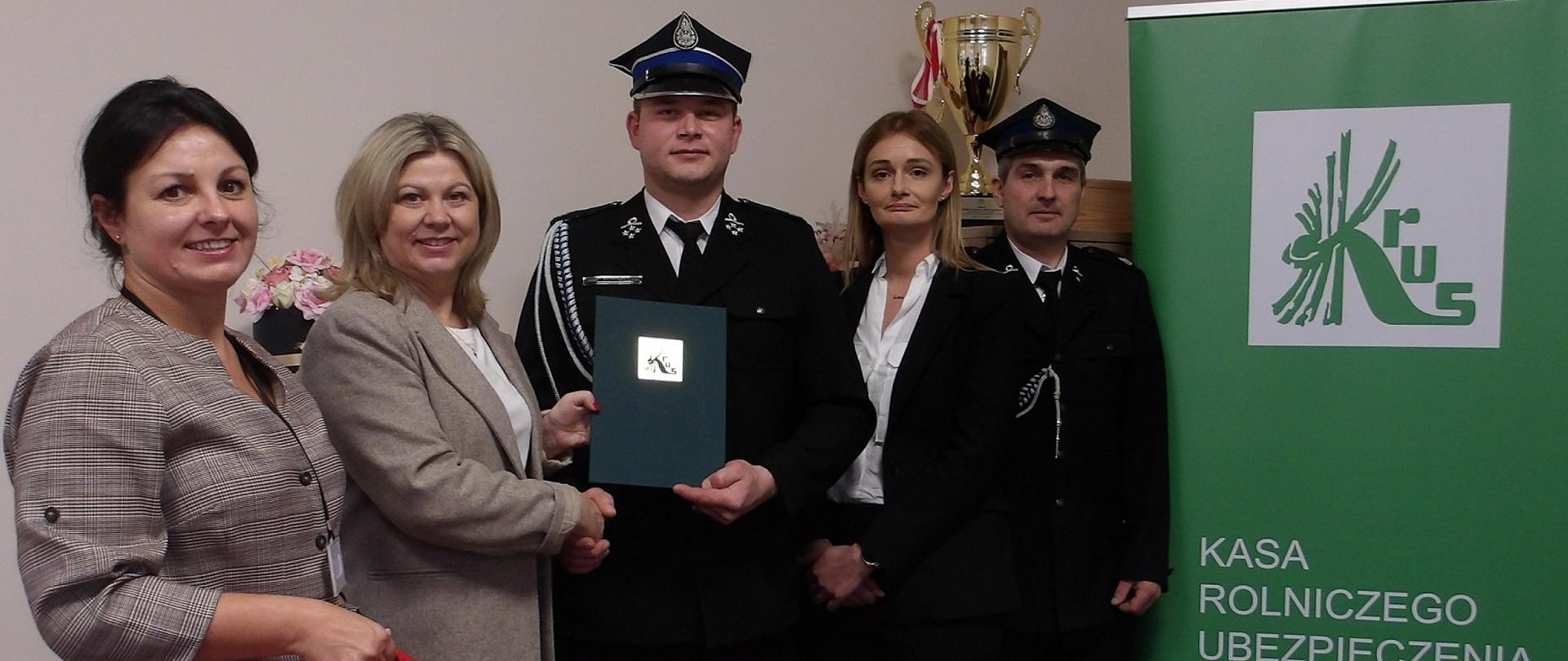 Pięć elegancko ubranych osób, w tym trzy kobiety i dwóch mężczyzn ubranych w mundury strażackie, pozuje w gabinecie. W tle złoty puchar i kwiaty, obok zielony roll-up w logo KRUS. Jedna z kobiet ściska dłoń strażakowi, wręczając mu jednocześnie zieloną teczkę z listem gratulacyjnym. Druga kobieta trzyma w ręce dużą czerwoną torbę ratowniczą z logo KRUS.