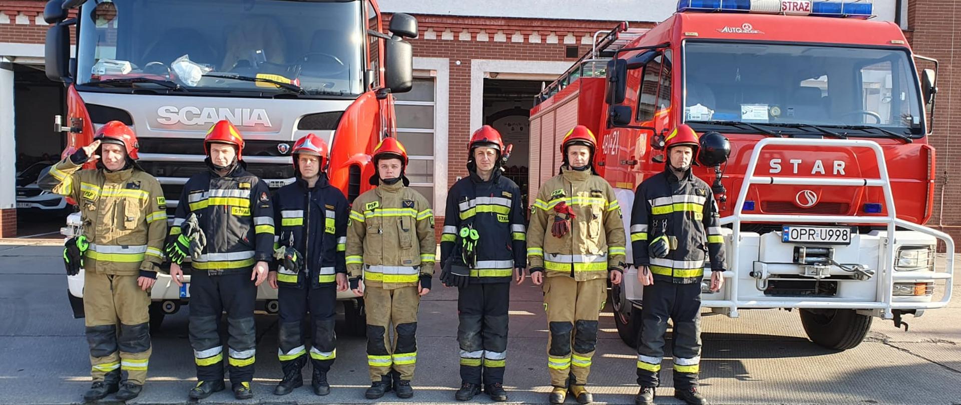 Zdjęcie przedstawia strażaków w mundurach bojowych podczas minuty ciszy dla poległych strażaków z Ukrainy na tle strażackich samochodów ratowniczo gaśniczych