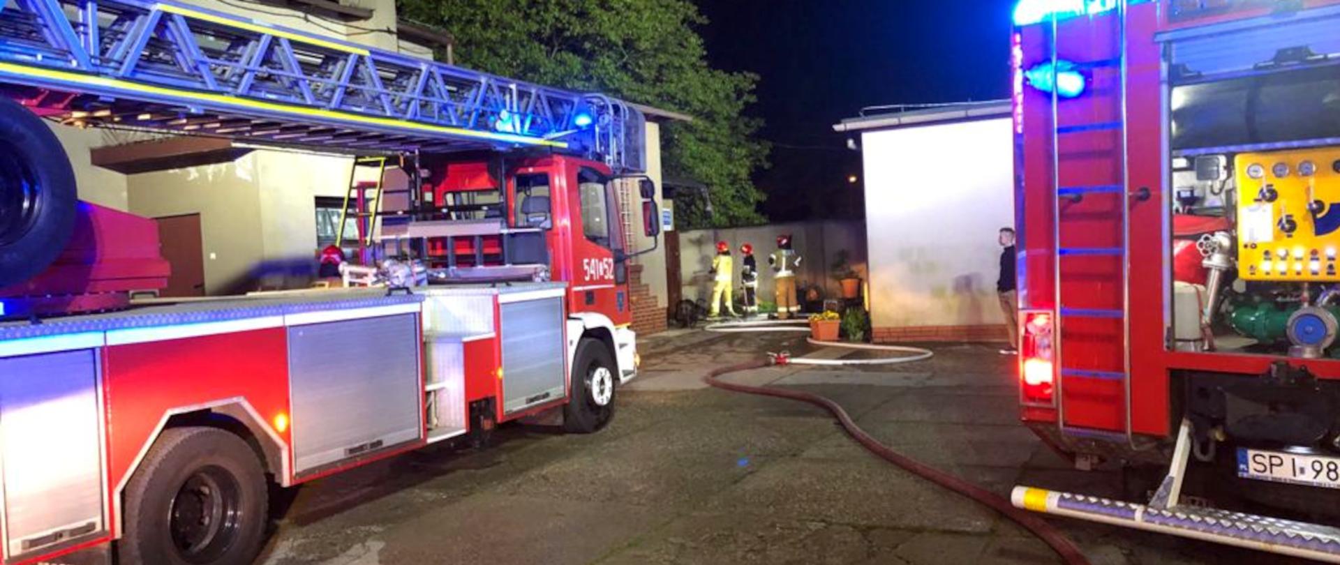 Zdjęcie przedstawia samochód specjalny drabina mechaniczna oraz średni samochód ratowniczo - gaśniczy oraz trzech strażaków w ubraniach specjalnych.