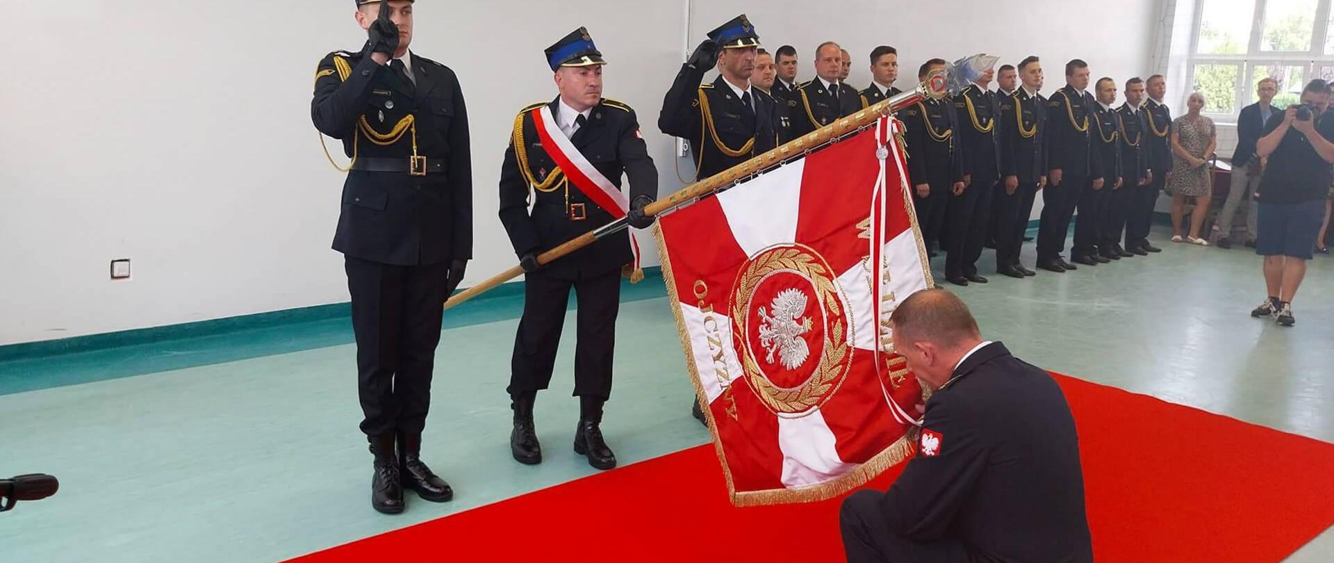 Na zdjęciu widać grupę osób strażaków w mundurach oraz osoby cywilne. Trzech strażaków w czapkach stoi przed czerwonym dywanem. Strażak w środku ma szarfę biało-czerwoną oraz trzyma sztandar. Naprzeciwko ich na czerwonym dywanie na jedno kolano klęka strażak bez czapki na głowie. Całuje sztandar, który jest przechylony w jego stronę. W tle widać pozostałe osoby. Uroczystość odbywa się na sali gimnastycznej. Jest widno