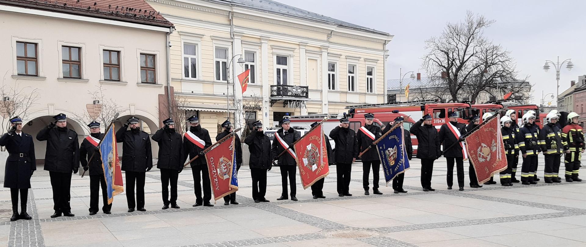 Na zdjęciu przedstawiono poczty sztandarowe ustawione na płycie Rynku. Od lewej: dowódca uroczystości kpt. Maciej Bartusiak, cztery poczty sztandarowe i druhowie ochotnicy z Ochotniczej Straży Pożarnej Niewachlów-Kielce. W tle zabudowania przy Rynku oraz samochody pożarnicze.