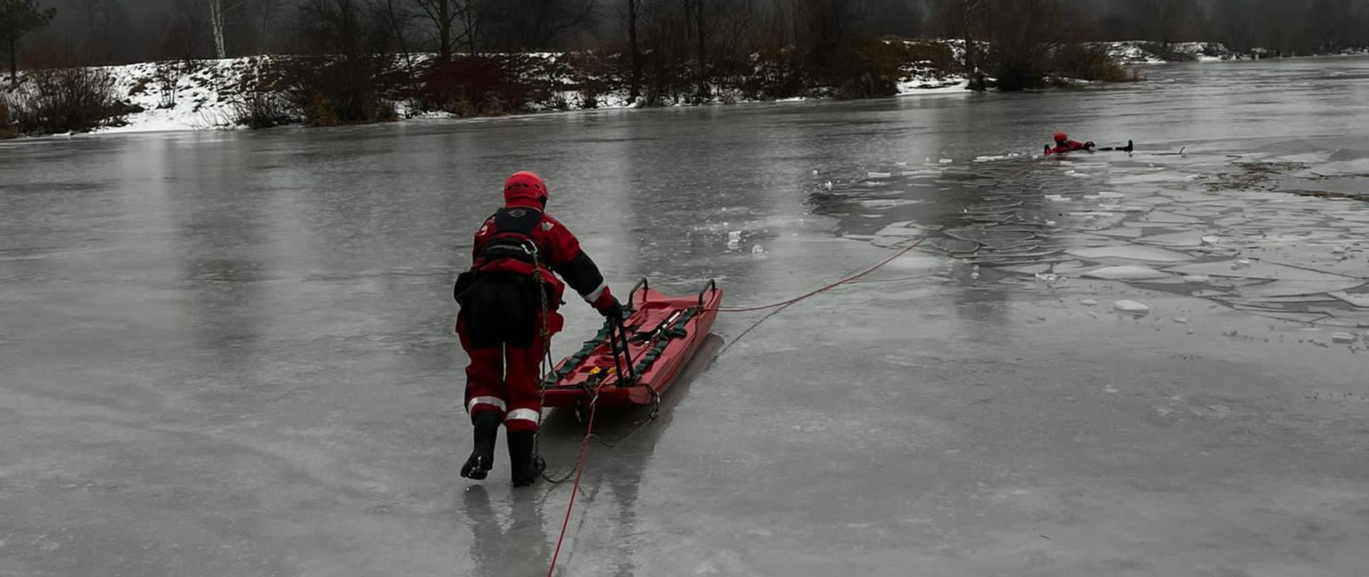 Zdjęcie przedstawia zalew w Mójczy i ratownika, który idzie z saniami lodowymi do osoby poszkodowanej znajdującej się w przerębli.