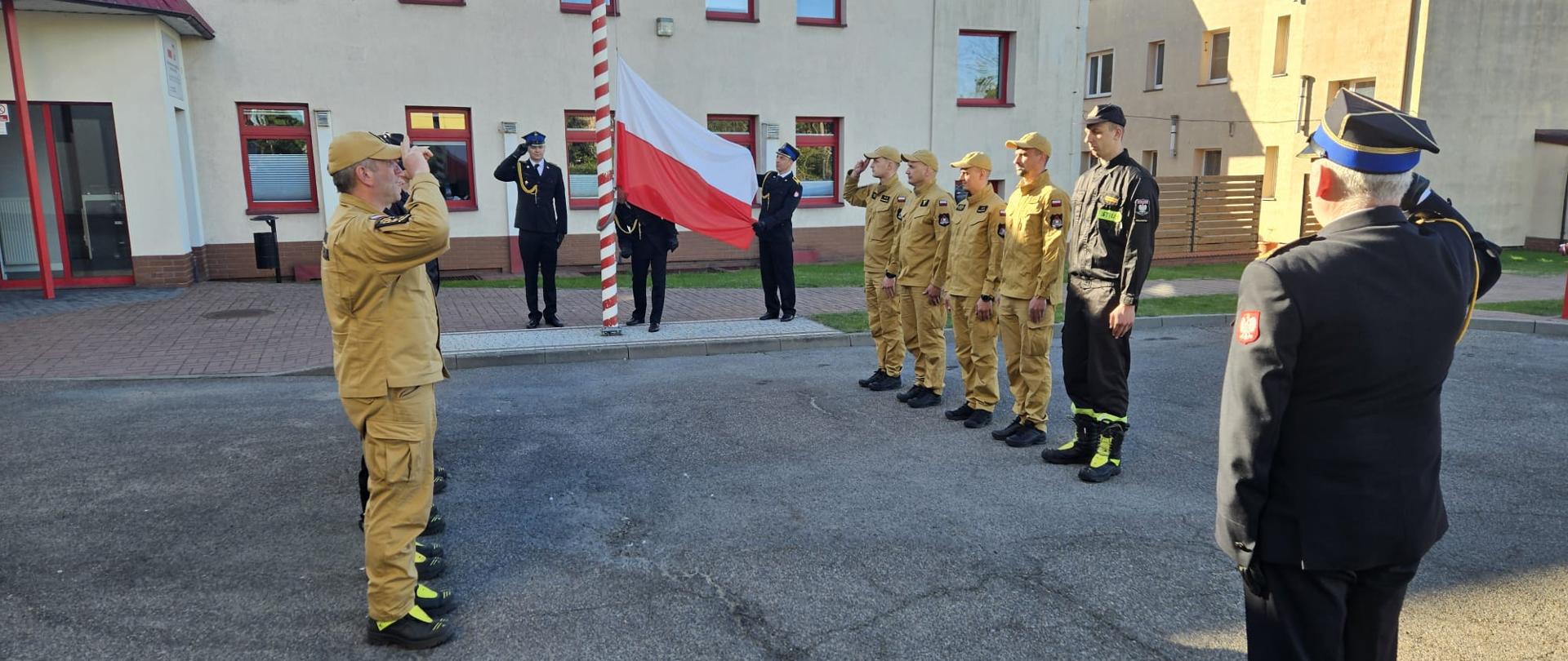 Uroczystość z okazji Dnia Flagi Rzeczypospolitej Polskiej. Strażacy ze zmian służbowych stoją naprzeciwko siebie. Poczet flagowy wciąga flagę państwową na maszt. Komendant, poczet flagowy dowódcy zmian salutują.