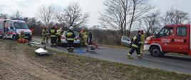 Zderzenie dwóch aut. Okolice Zaborowic. Odcinek jezdni, na którym doszło do wypadku. Strażacy wykonują dostęp do osób znajdujących się w volkswagenie stojącym na środku drogi. Przed autem znajduje się sprzęt hydrauliczny. Po prawej stronie wóz straży pożarnej, po lewej - karetka pogotowia ratunkowego. W tle drzewa.