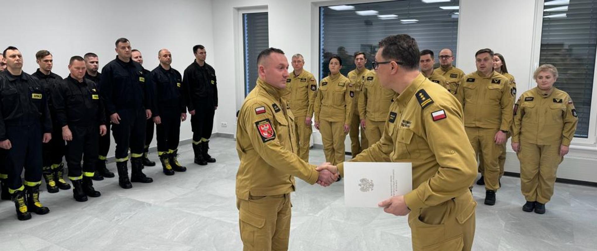 Na sali szkoleniowej stoją w mundurach strażacy. Na środku sali komendant miejski ściska rękę awansowanemu strażakowi i wręcza mu teczkę z dokumentami.