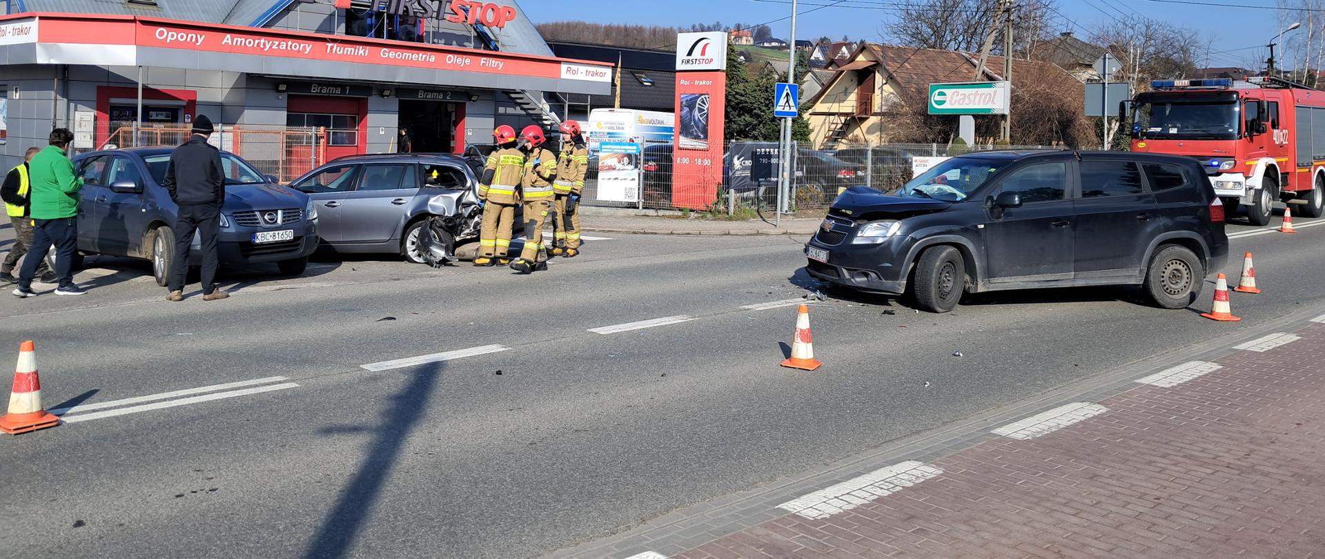Zdjęcie przedstawia zderzenie trzech samochodów osobowych. Na zdjęciu widać na środku jezdni stoi samochód osobowy z uszkodzonym przodem. Po lewej stronie widać dwa pojazdy jeden z nich ma uszkodzony tył a drugi stoi przodem. Obok tych samochodów stoi trzech strażaków ubranych w ubrania specjalne w kolorze piankowym na głowie mają czerwone hełmy. Przed pojazdami na drodze widać rozstawione pachołki ostrzegawcze. Po prawej stronie stoi czerwony pojazd pożarniczy. Za pojazdami widać budynek warsztatu samochodowego. Po lewej stronie stoją osoby postronne