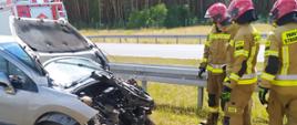 Kolizja na drodze ekspresowej. Jezdnia w kierunku Poznania. Na pasie zieleni stoi samochód osobowy uczestniczący w kolizji. Auto ma bardzo mocno uszkodzony przód. Obok samochodu znajduje się 3 strażaków. W tle las.
