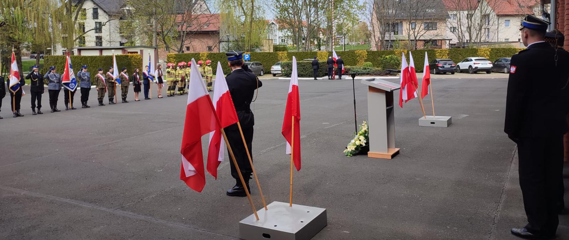 Na placu wewnętrznym strażackim biało czerwone flag w tle poczty sztandarowe oraz drzewa.
