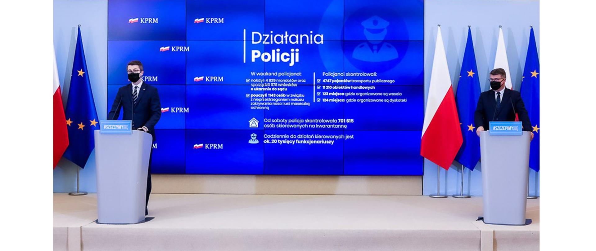 Na zdjęciu widać stojących za mównicami Piotra Mullera, rzecznika rządu i Macieja Wąsika wiceministra MSWiA. W tle widać ekran i flagi Polski i UE.