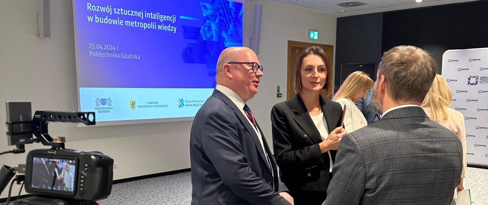 sala, w tle napis wyświetlony na tablicy „Rozwój sztucznej inteligencji w budowie metropolii wiedzy", stoją trzy osoby (w tym wojewoda) i dyskutują